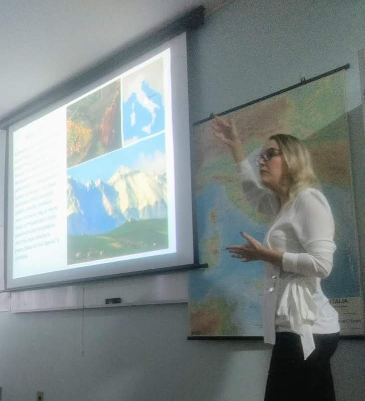 Professora em aula sobre a Itália, ao lado de uma apresentação e de um mapa